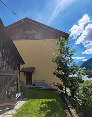 Un bâtiment doté d’un toit en pente, d’une petite structure adjacente avec une porte, et d’une cour verdoyante avec un banc.
