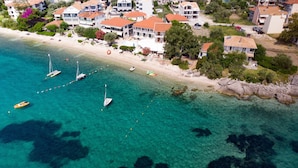 An aerial view of a coastal area with clear turquoise waters, several sailboats anchored near the shore, and a row of buildings with red-tiled roofs.