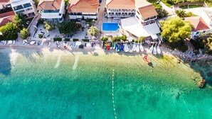 An aerial view of a coastal hotel with clear waters, a swimming pool, and a beach area with sun loungers.