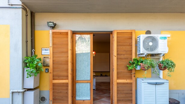Entrada de un hotel con contraventanas de madera, un equipo de aire acondicionado y plantas en macetas.