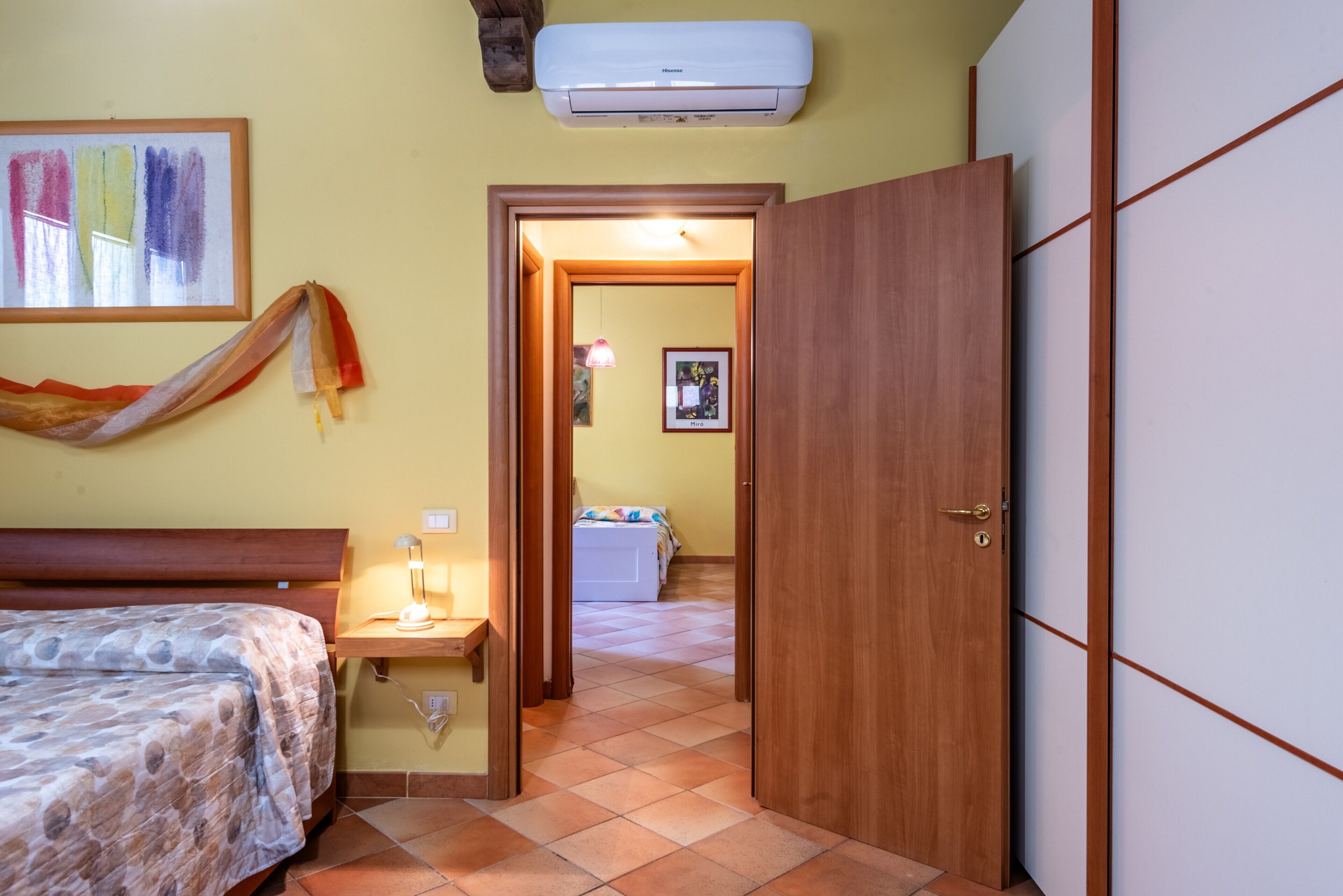 A bedroom with a bed, bedside table, lamp, air conditioning unit, and a door leading to another room.