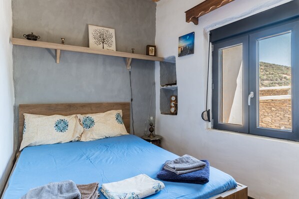A bedroom with a bed, a window with a view, a shelf with decorative items, and a bedside table with a lamp.