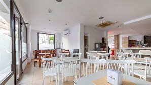 A modern dining area with white tables and chairs, a kitchen in the background, and a window with a view of palm trees.