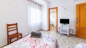 A bedroom with a bed, a wooden chair, a television on a white stand, a wooden door, and a bathroom visible through an open door.