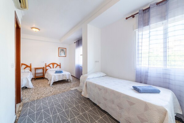 A bedroom with two single beds, a small table, a chair, a patterned rug, and a window with sheer curtains.