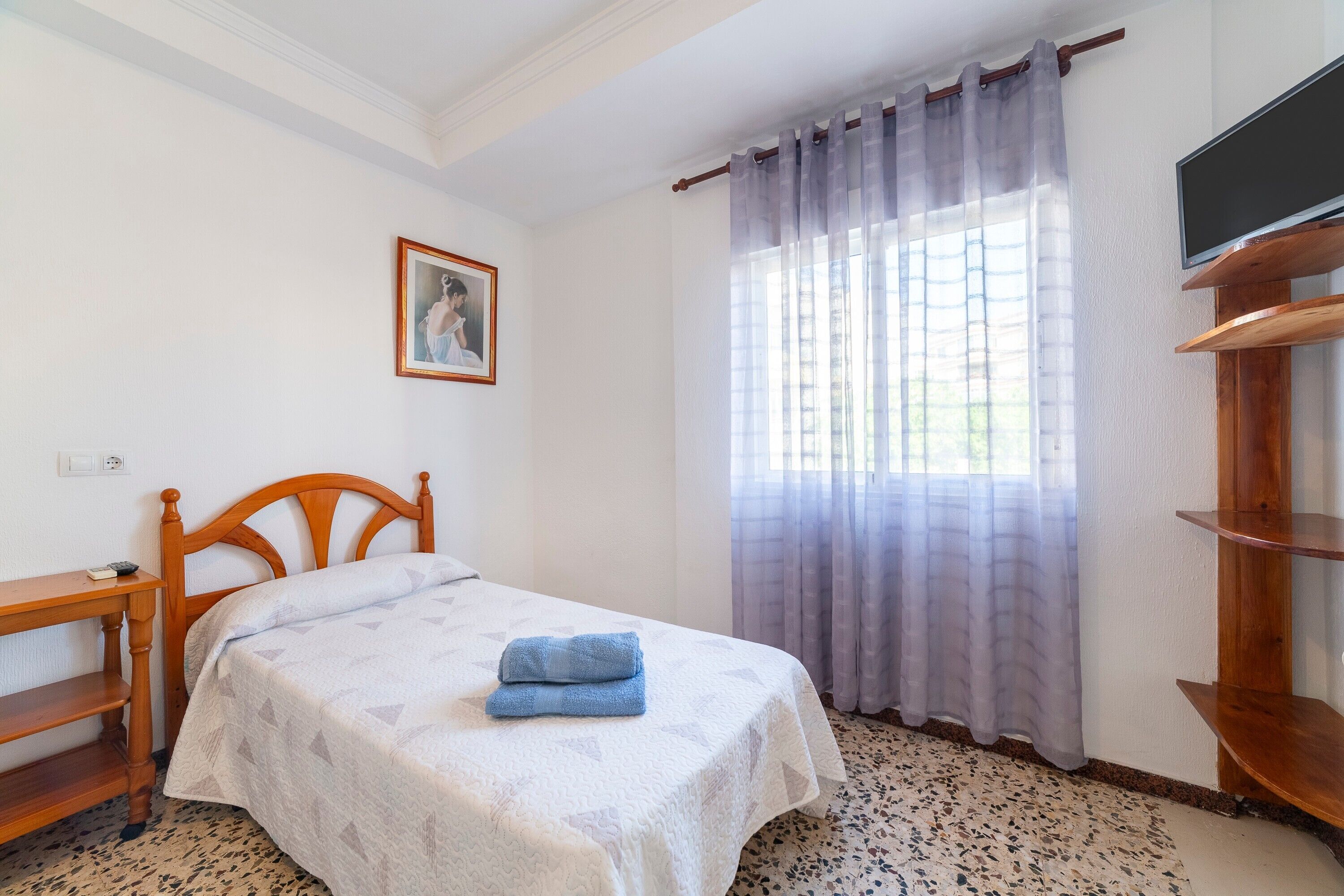 A bedroom with a bed, a wooden headboard, a bedside table, a TV on a wooden stand, a window with sheer curtains, and a framed picture on the wall.