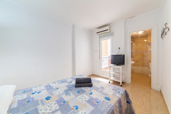 A bedroom with a bed, a TV on a stand, an air conditioning unit, and a bathroom visible through an open door.