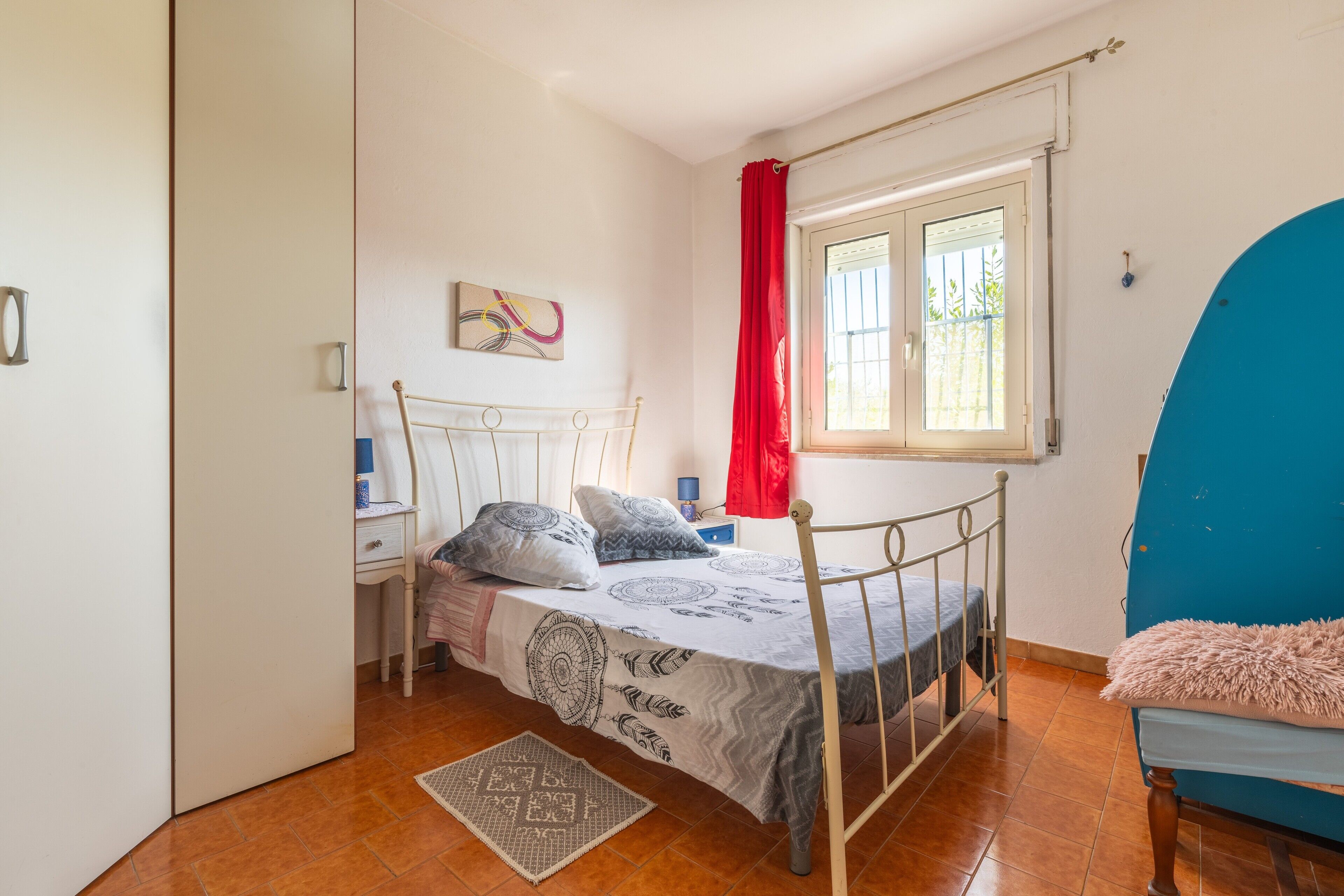 Ein Schlafzimmer mit einem Bett, einem Fenster mit Vorhängen, einem Nachttisch und einem Sessel.