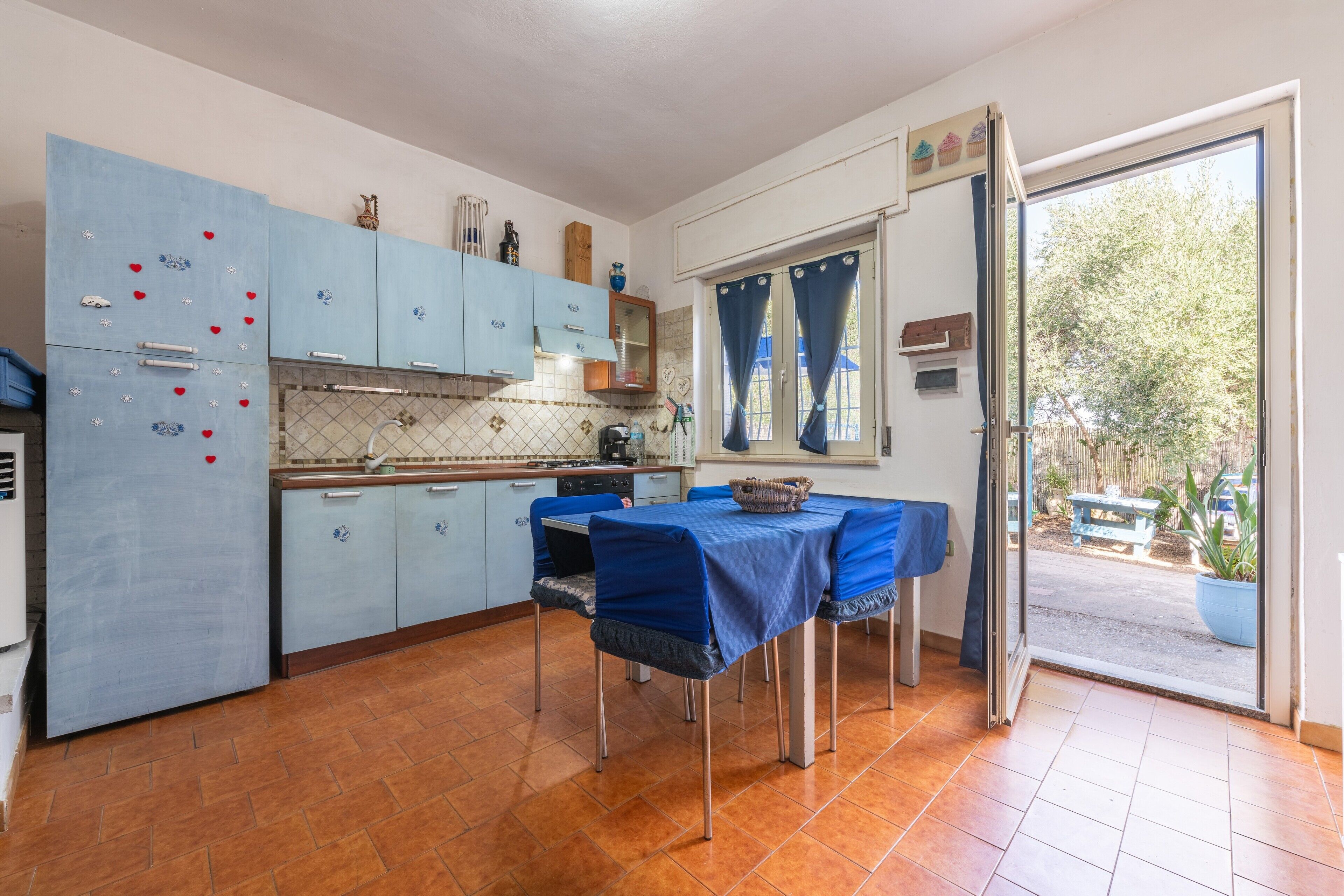 Une cuisine avec des meubles de rangement bleus, une table à manger recouverte d’une nappe bleue et une vue sur l’extérieur.