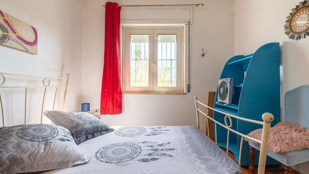 A bedroom with a bed, a window with curtains, a blue chair, and a wall-mounted fan.