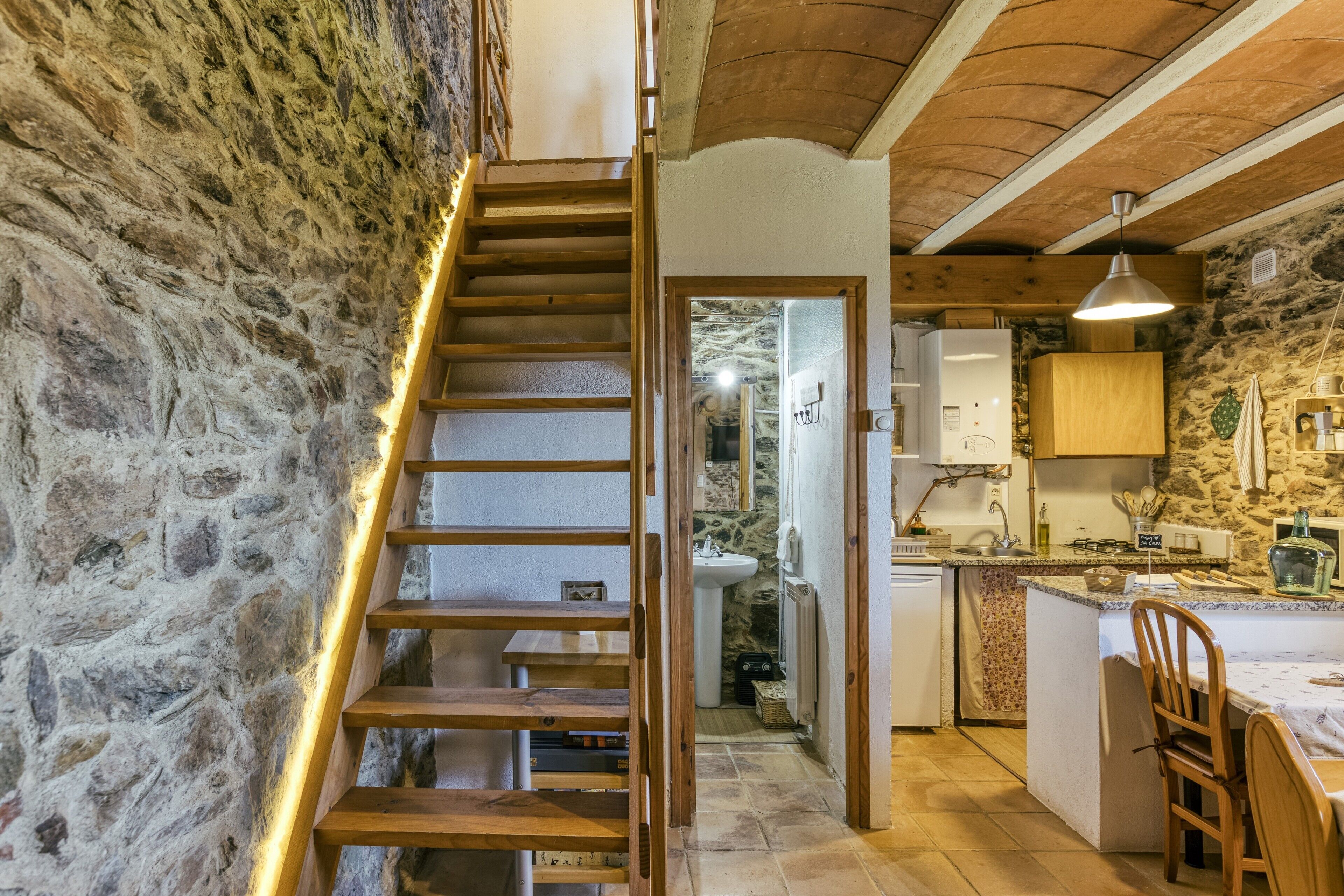 Una cocina rústica con pared de piedra, escalera de madera y una pequeña isla con pileta y encimera.