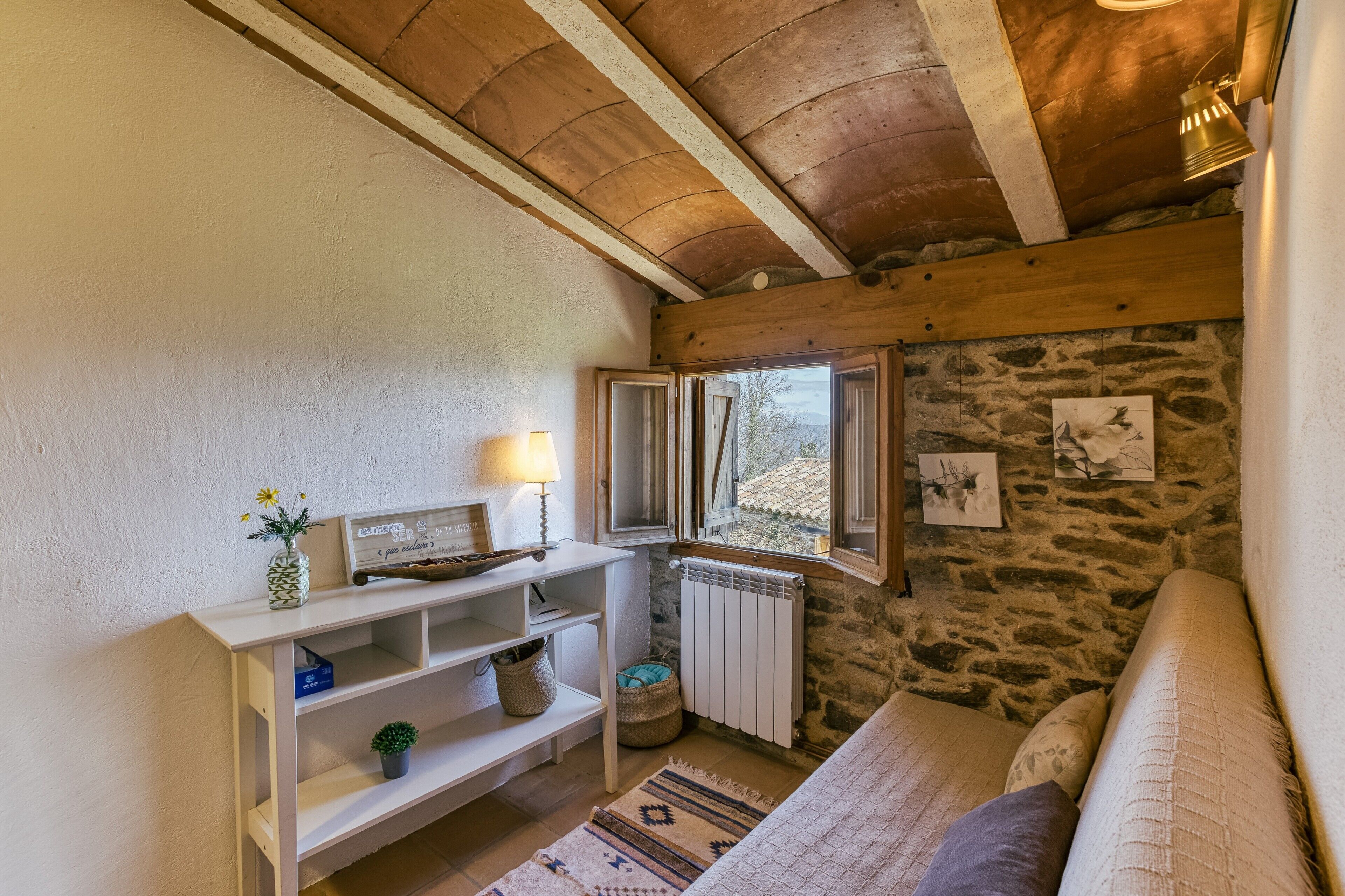 Una habitación acogedora con pared de piedra, vigas de madera, un sofá blanco, una mesita con una lámpara y una ventana con vistas.