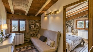 A cozy bedroom with a stone wall, wooden ceiling beams, a bed, a sofa, and a view of the outdoors. - Apartment 'La Rústica' with Mountain View, Shared Garden and Wi-Fi (susqueda)