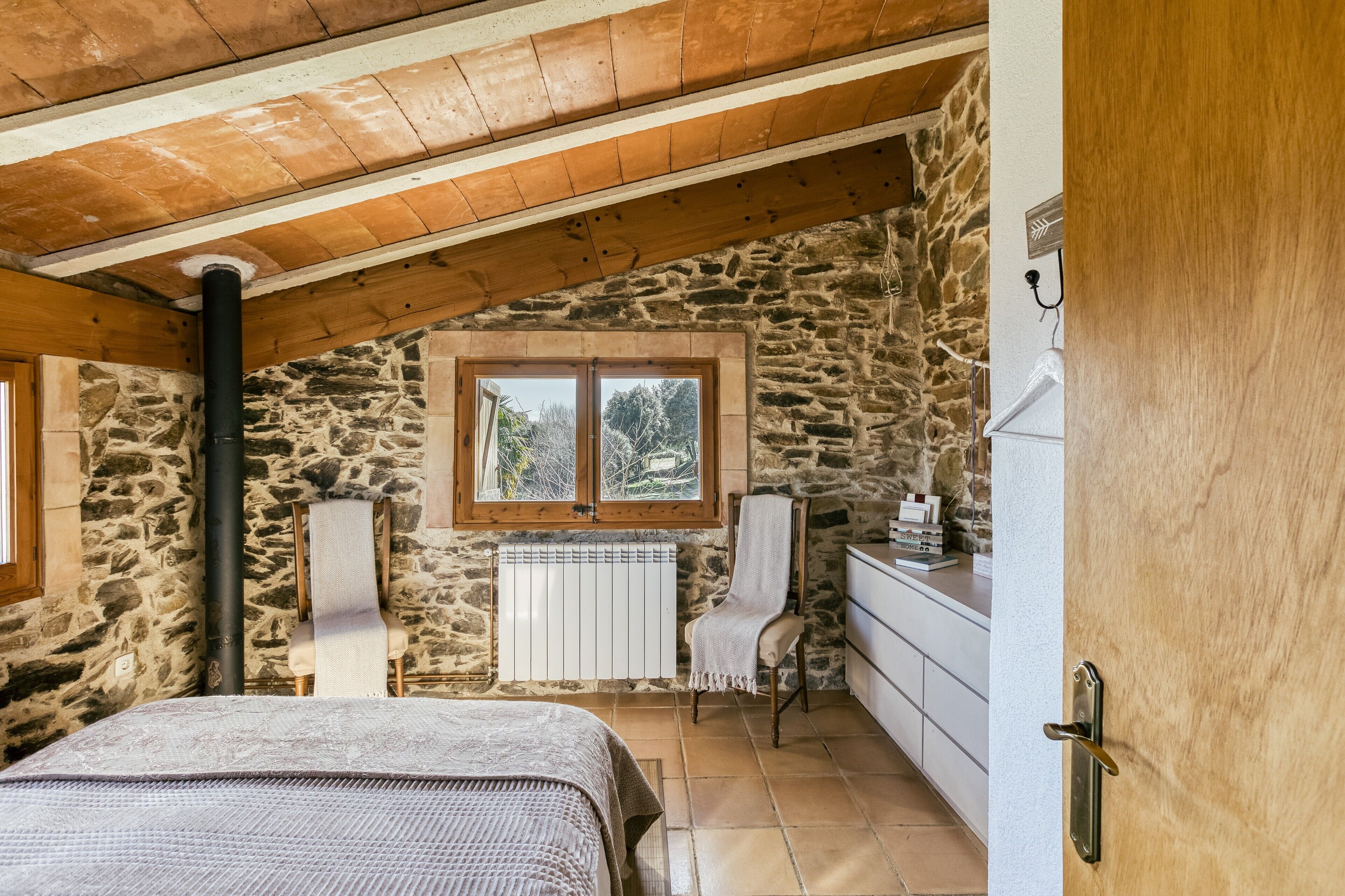 A bedroom with a stone wall, a wooden ceiling, a bed, a chair, a window, and a radiator.