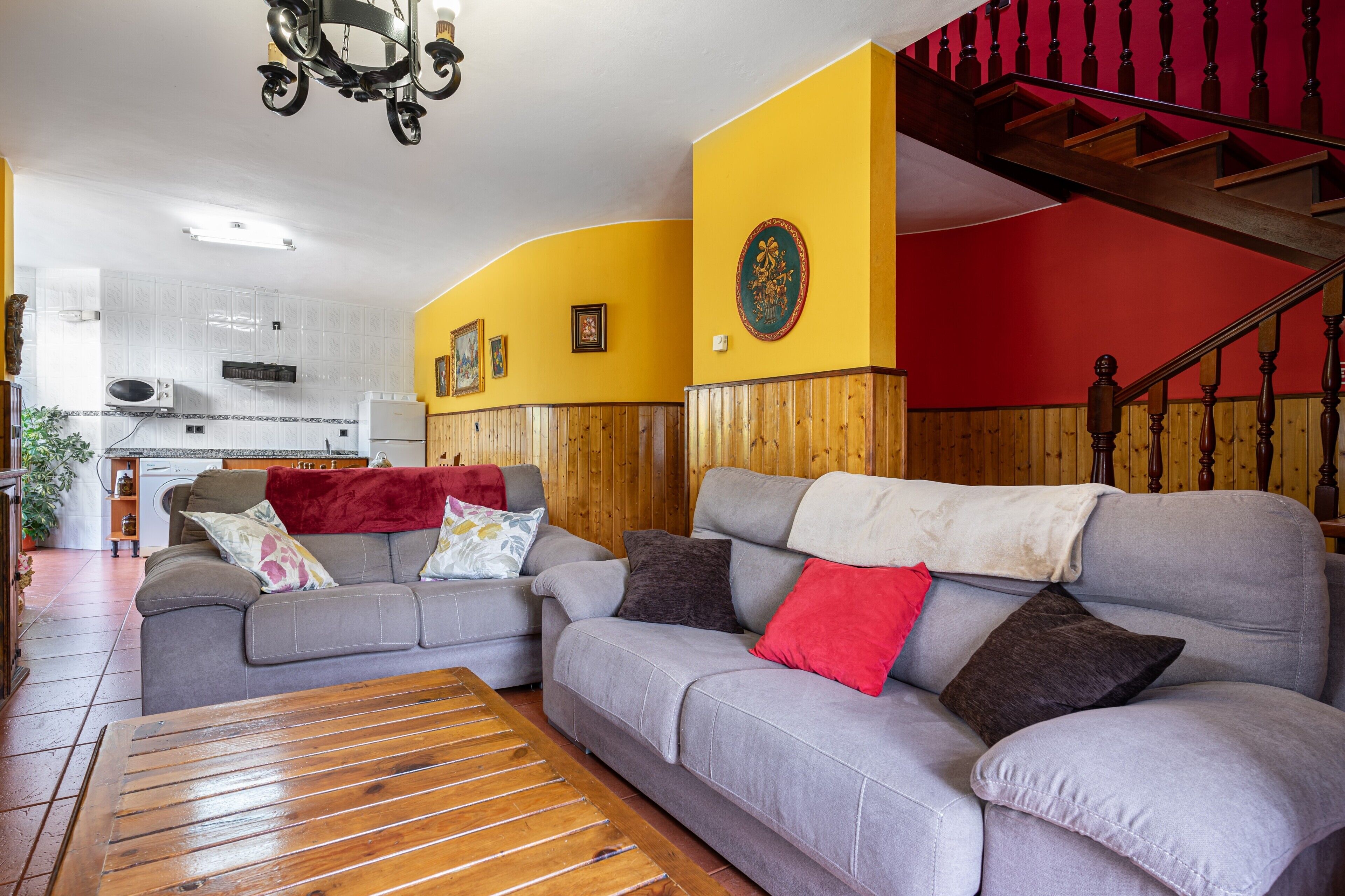 Un salon avec deux canapés, une table basse en bois, un escalier avec des rampes en bois, et une cuisine à l’arrière-plan.