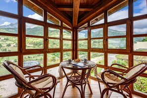 Una habitación con ventanas amplias, sillas de mimbre, una mesa con un libro y vistas a un paisaje montañoso.