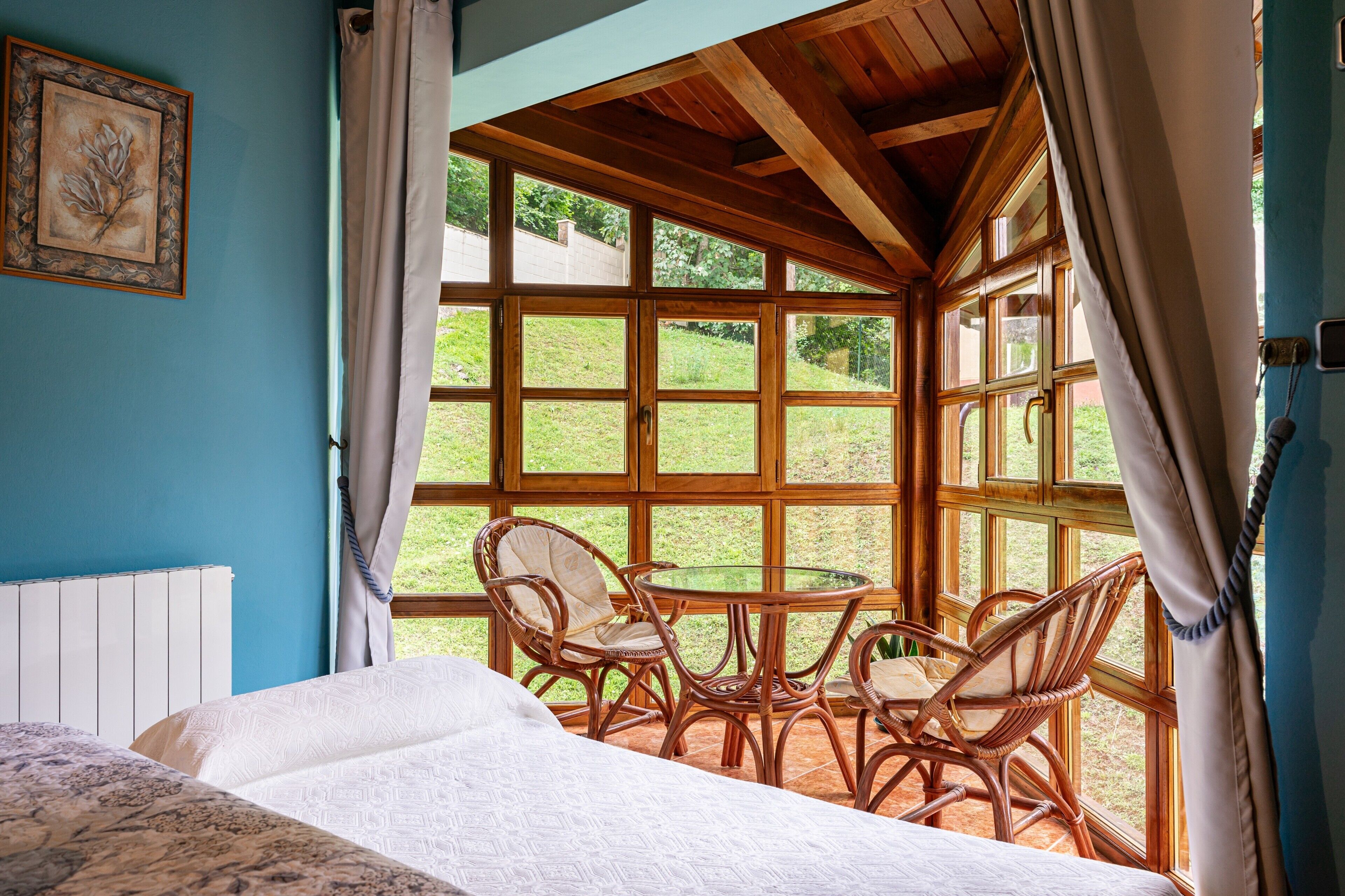 Un dormitorio con cama, ventana con cortinas, sillas de mimbre y vistas al verdor.