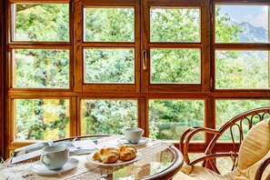 A room with a table set for breakfast, a wicker chair, and a view of greenery through large windows.