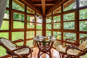 Una habitación con ventanas amplias, sillas de mimbre, una mesa redonda con mantel blanco y una planta verde.