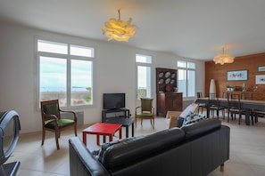 A living room with a sofa, chairs, a TV, a dining table, and a kitchen area with a wooden wall and a view.