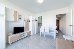 Una sala de estar moderna y luminosa con un televisor de pantalla plana, muebles blancos y una cocina al fondo.