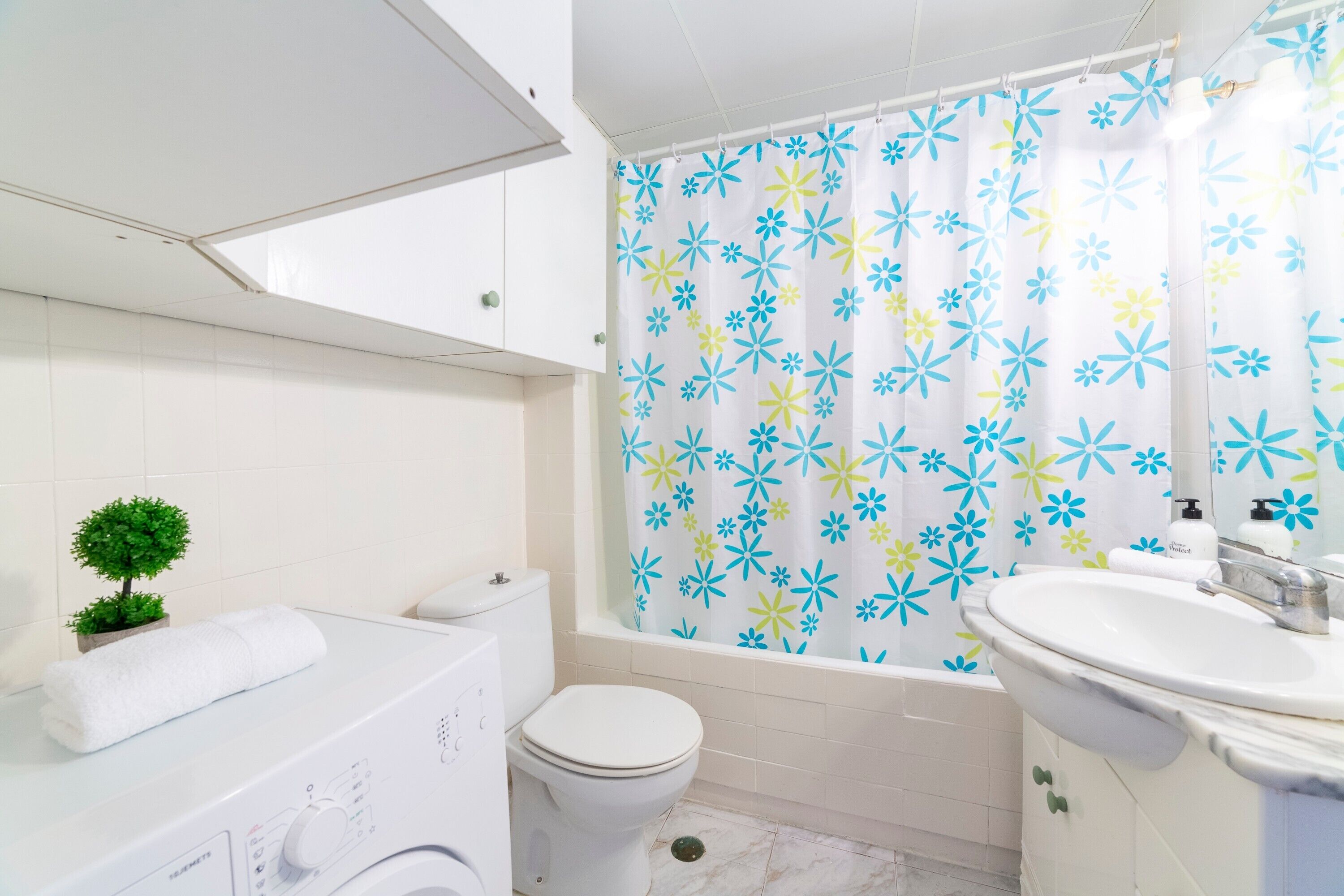 A bathroom with a white toilet, a white sink, a bathtub with a floral shower curtain, and a small potted plant.
