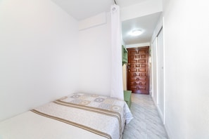 Un pasillo estrecho que conduce a una puerta de madera, una cama con ropa de cama estampada y una caja verde en el suelo.