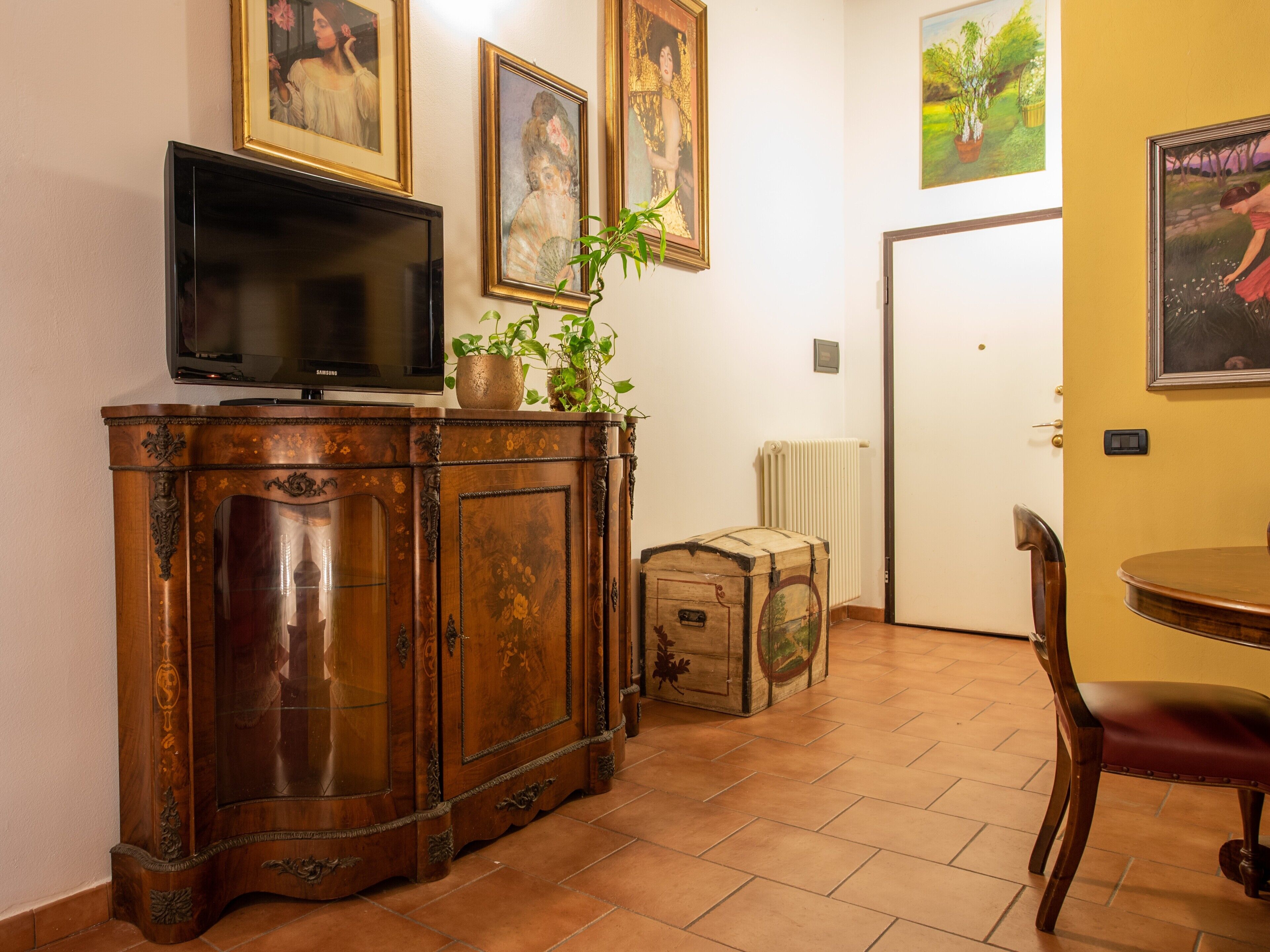 A room with a wooden cabinet, a television, a potted plant, a suitcase, and a dining table with a chair.