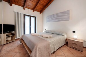 A bedroom with a bed, a TV, a bedside table with a lamp, a radiator, and a window.
