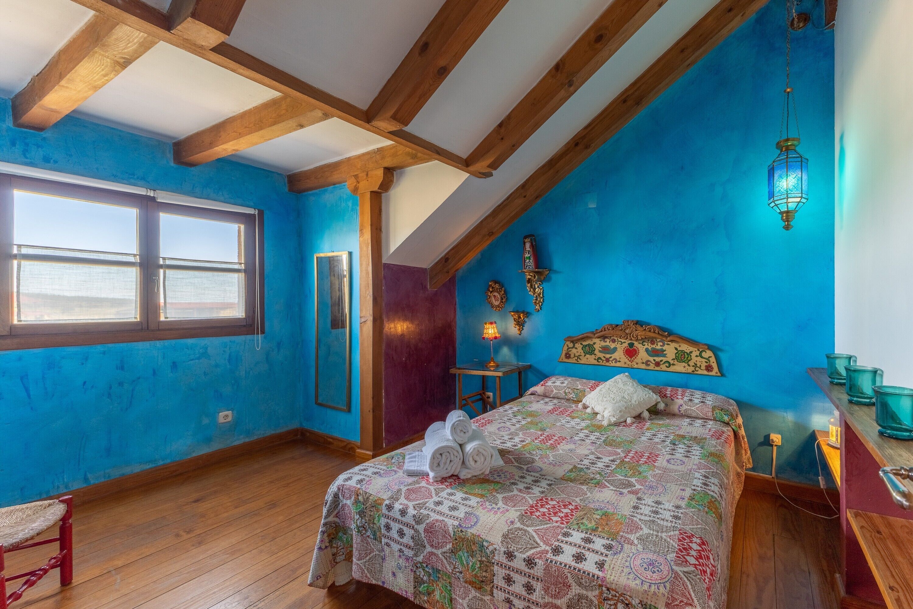Een slaapkamer met een schuine kap, houten balken, een groot bed met een beddeken met dessin, een raam en een klein tafeltje met een lamp.