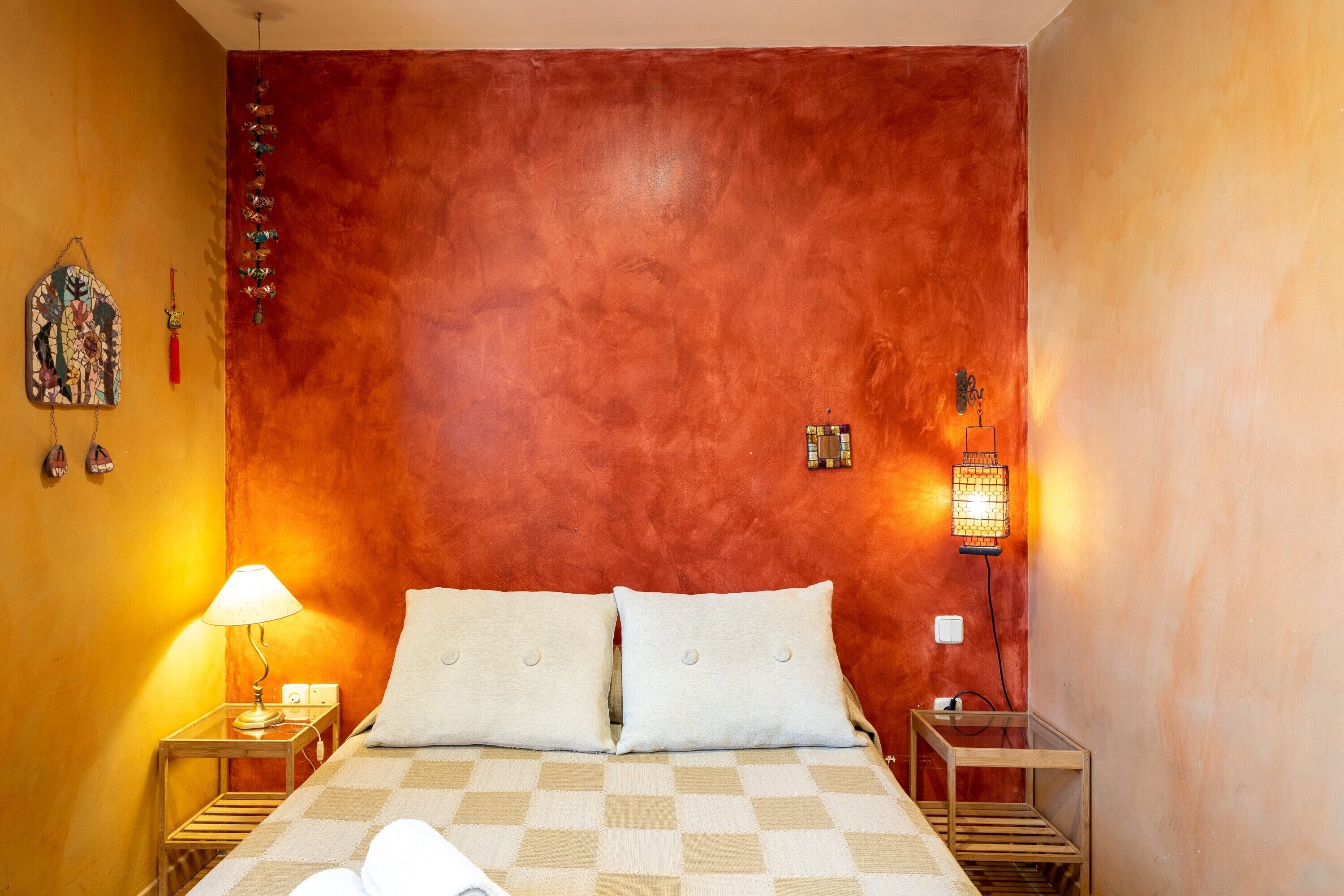 A room with a checkered bedspread, two pillows, a nightstand with a lamp, a wall-mounted light, and a small table with a lamp.