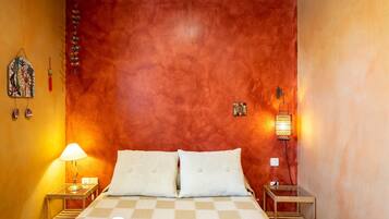 A room with a checkered bedspread, two pillows, a nightstand with a lamp, a wall-mounted light, and a small table with a lamp.