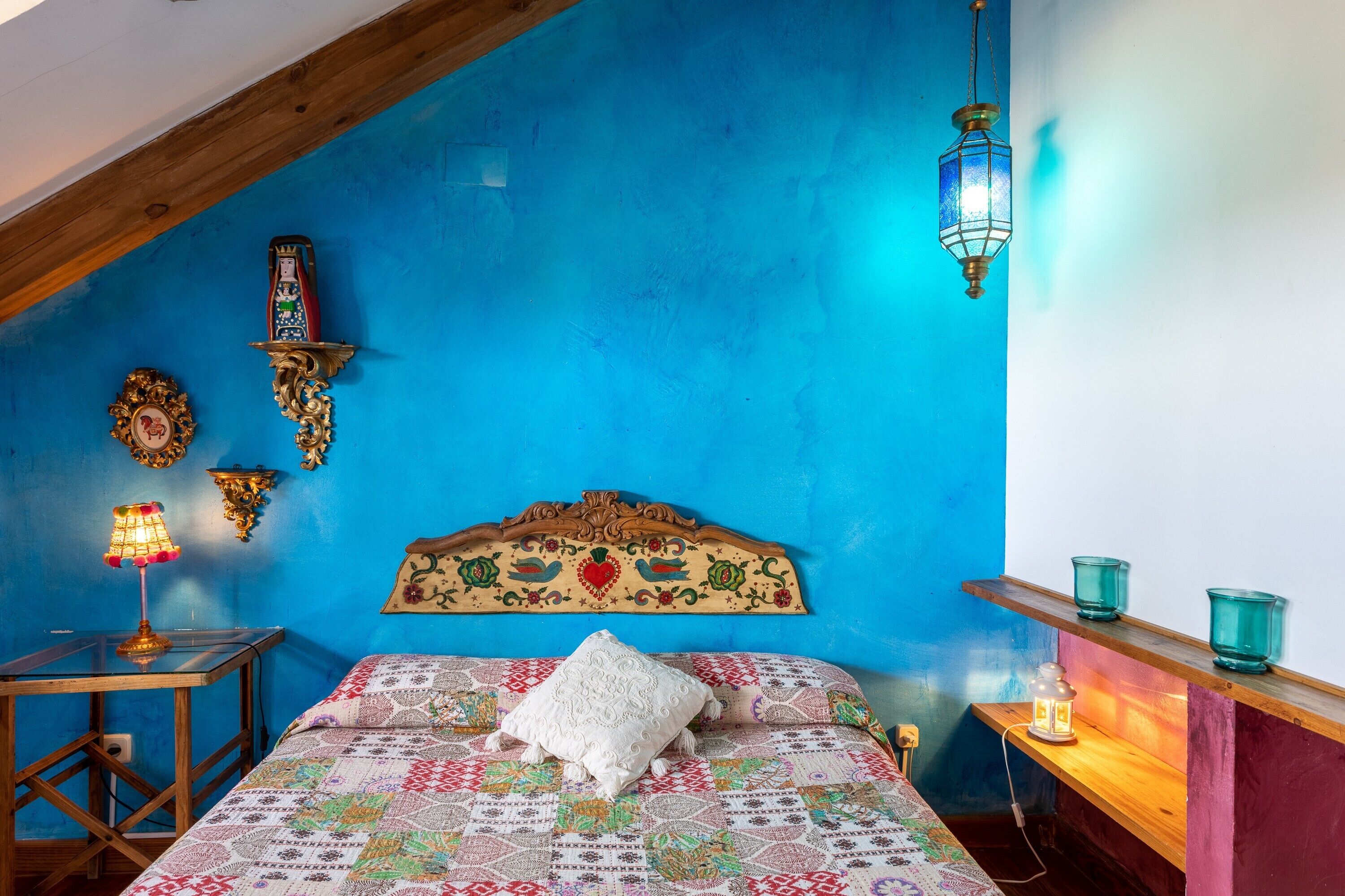 Un dormitorio con pared azul, cabecera de madera, colcha estampada, almohada blanca, mesita de noche con lámpara y apliques de pared.