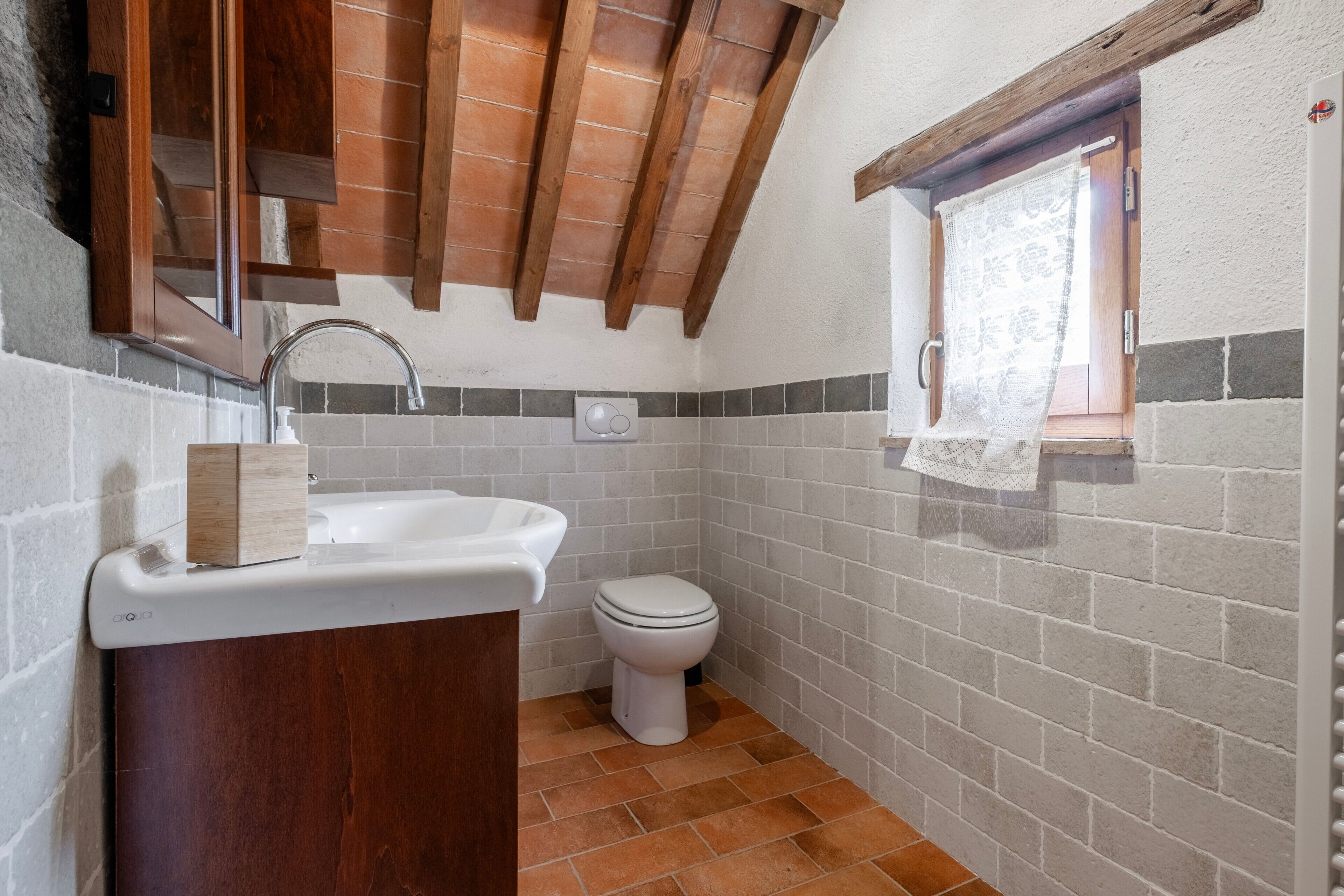 A compact bathroom with a wooden vanity, a white sink, a toilet, and a small window with a curtain.
