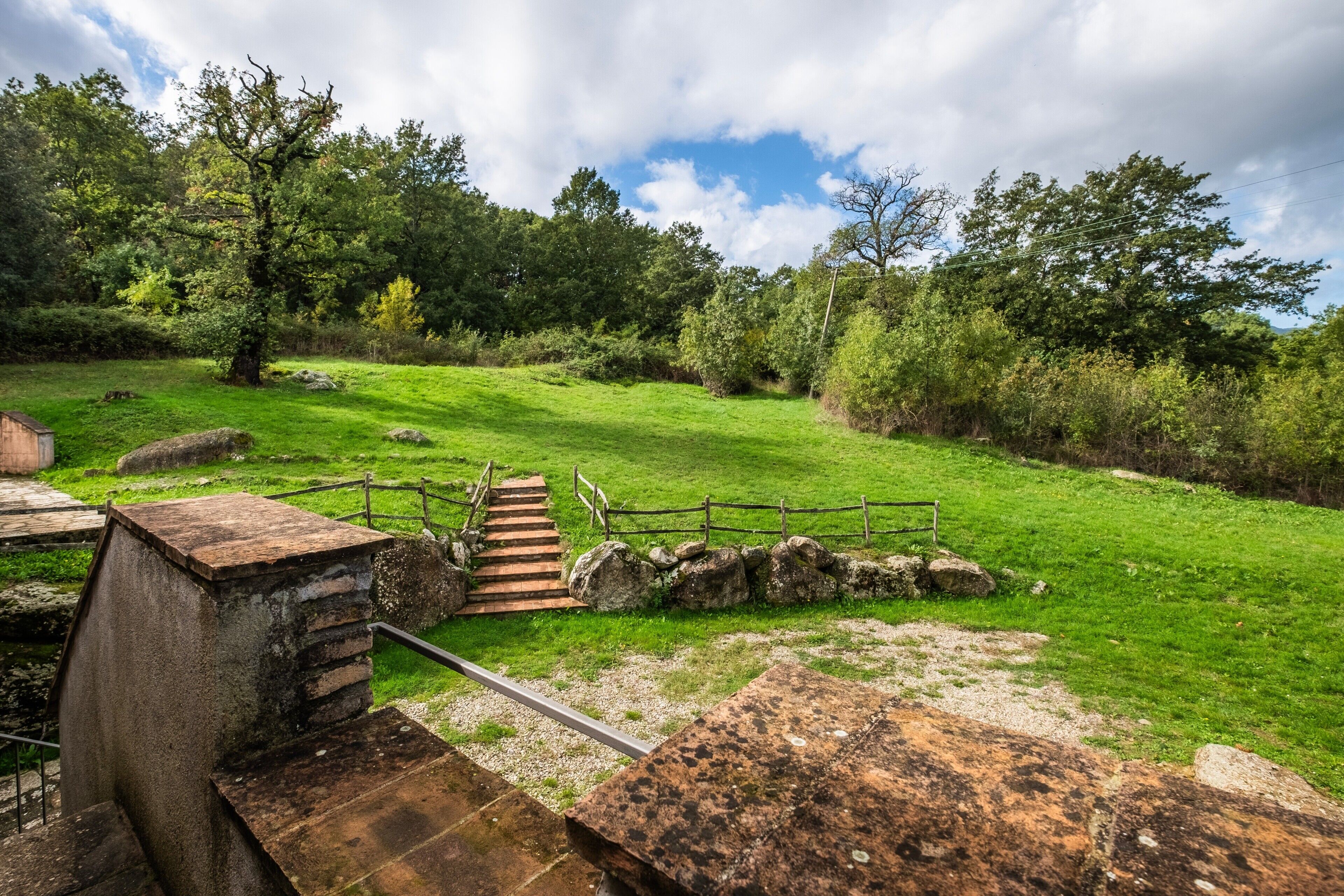 Un escalier en pierre rustique mène à une terrasse en bois, qui est entourée d’une végétation luxuriante et s’ouvre sur un ciel dégagé.