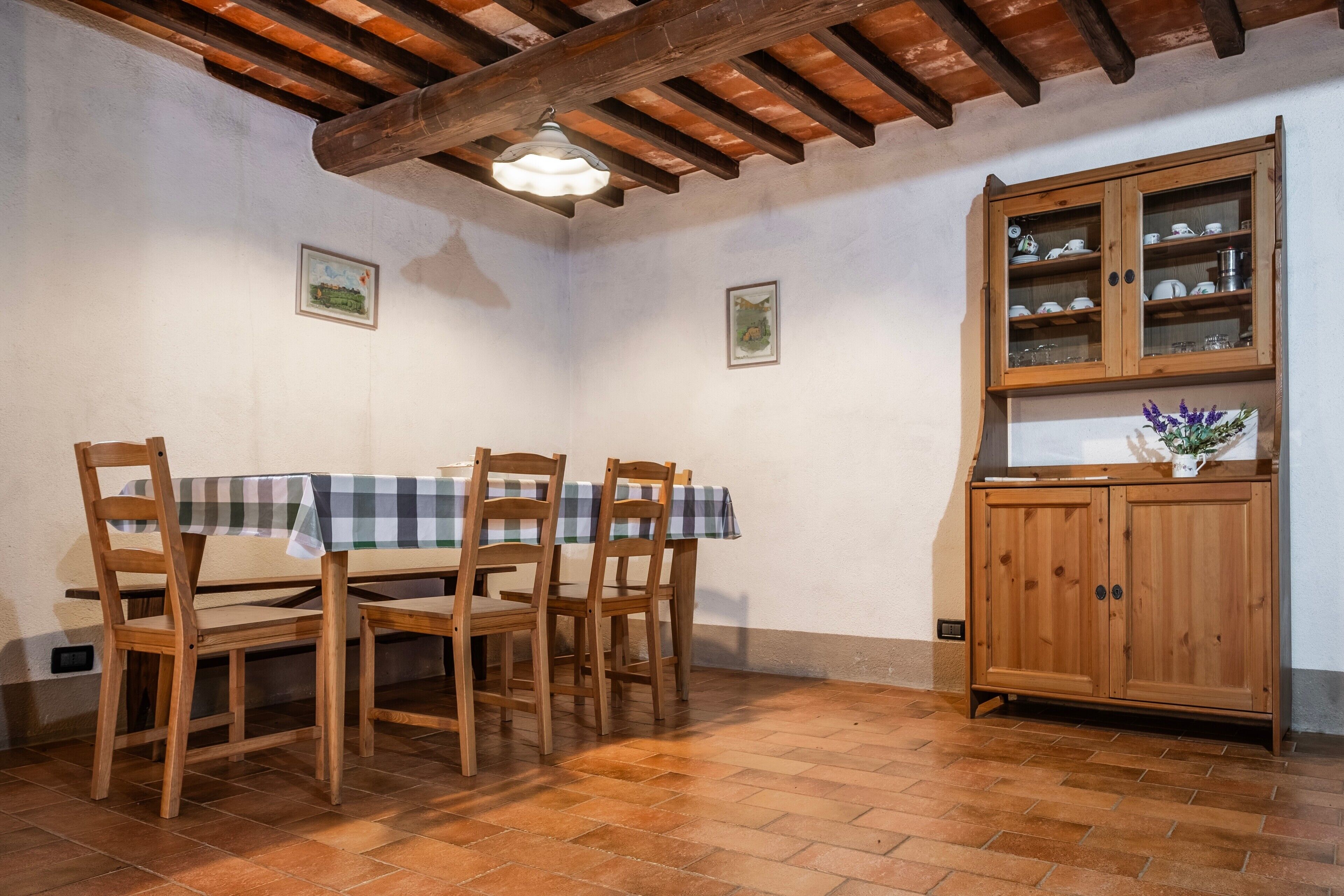 Uma sala de jantar com mesa e cadeiras de madeira, um armário com portas de vidro e chão ladrilhado.