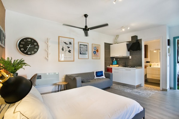 A modern hotel room with a bed, a sofa, a small table, a clock, a ceiling fan, and a kitchenette.