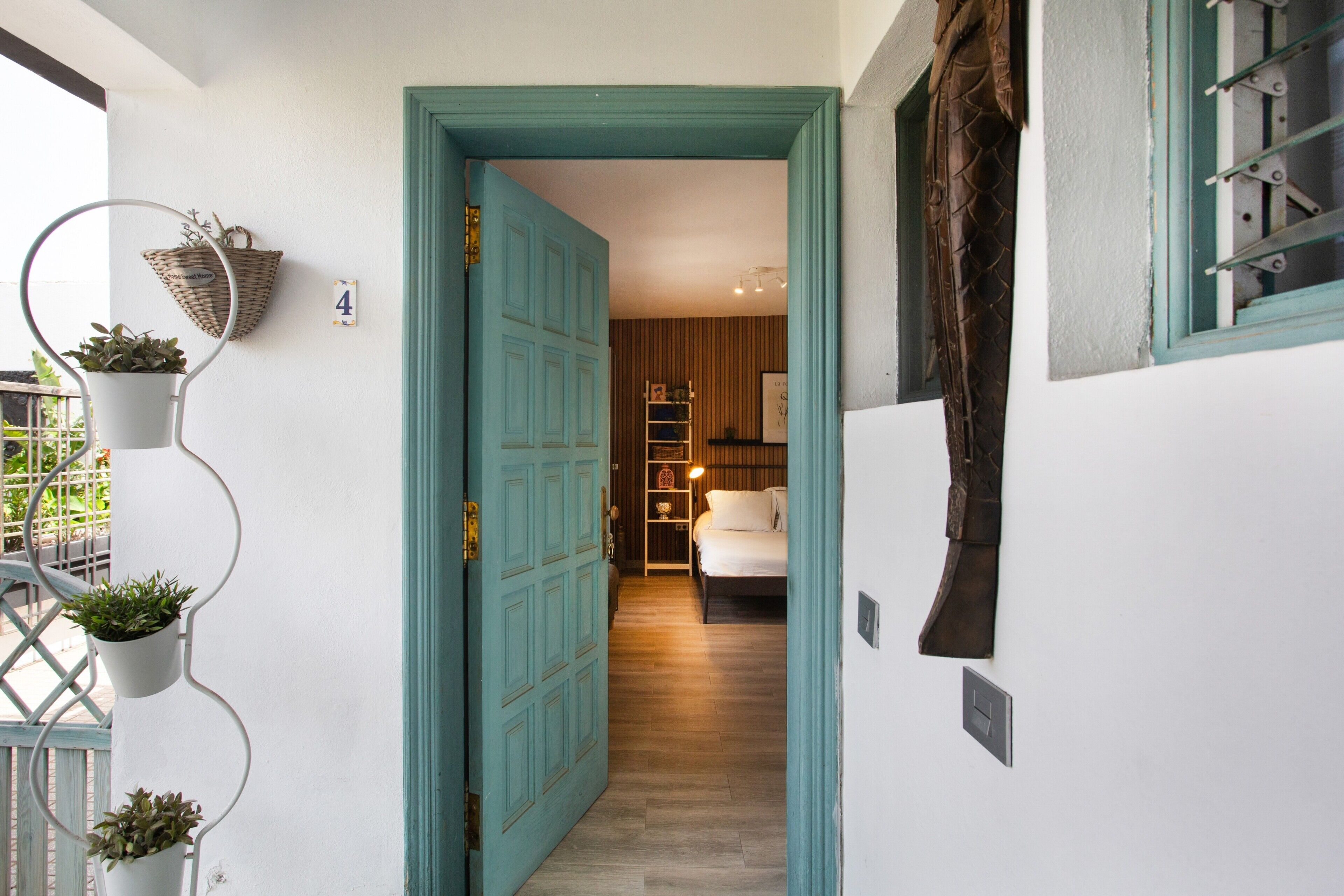 A hallway with a teal door leading to a bedroom with a bed and a ladder. There are two hanging planters on the left.