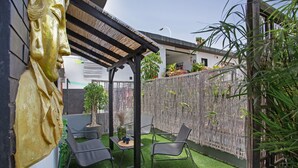 Un patio exterior con césped artificial, dos sillones de descanso, una mesa con un florero y una estatua dorada decorativa.