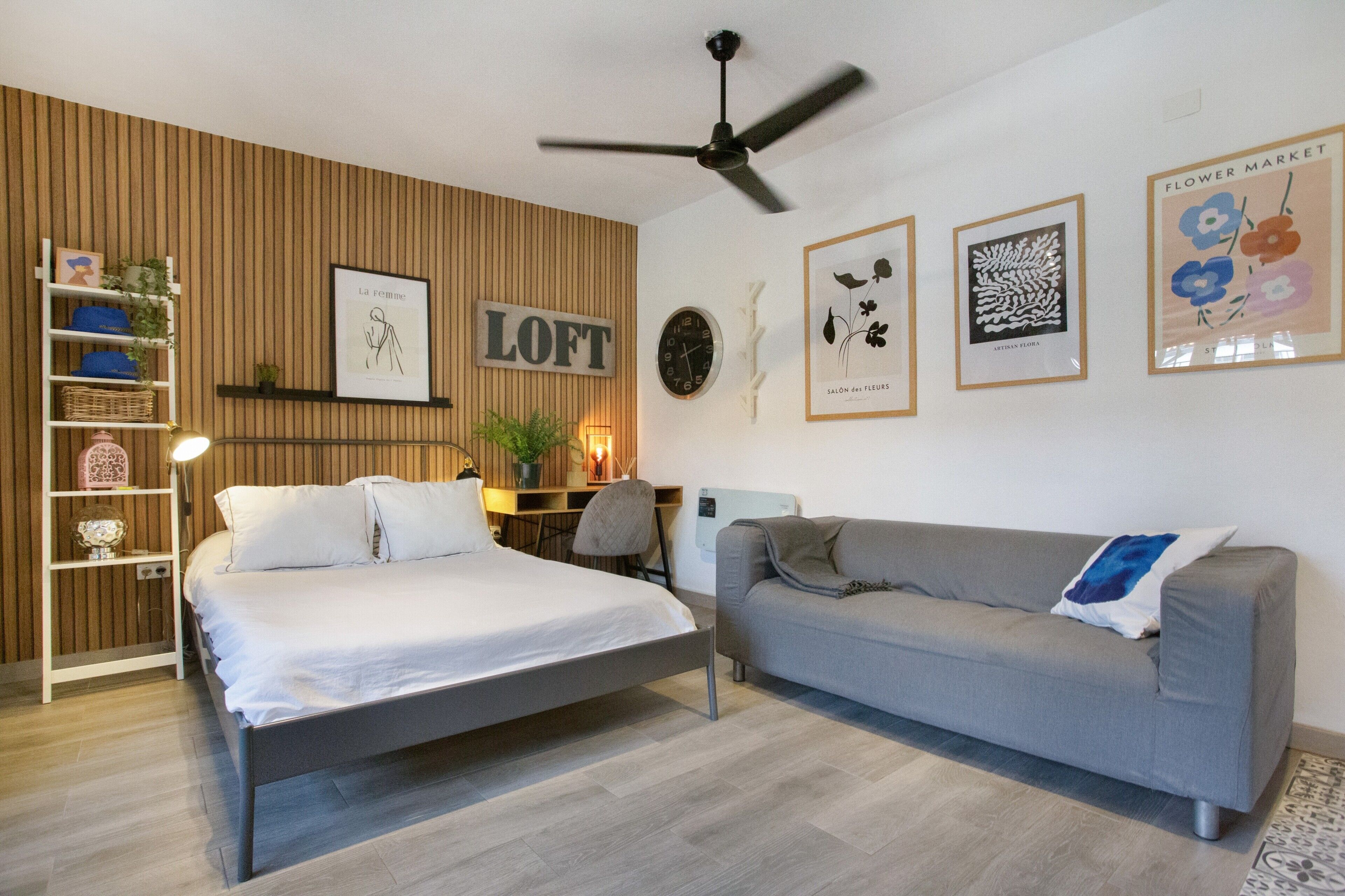 Een slaapkamer met een bed, een grijze bank, een houten wandpaneel, een plafondventilator en kunstwerken aan de muur.