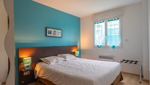 Quarto com cama, mesas de cabeceira, um quadro na parede, janela com cortinas e radiador.