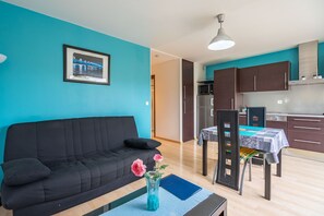 A living room with a black sofa, a dining table with chairs, a vase with flowers, and a kitchen in the background.