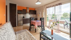 Cozinha moderna com armários escuros, micro-ondas e geladeira. Há uma mesa de jantar com toalha quadriculada vermelha e branca e cadeiras. Um sofá está localizado na área de estar, e uma janela com cortinas leves é visível.