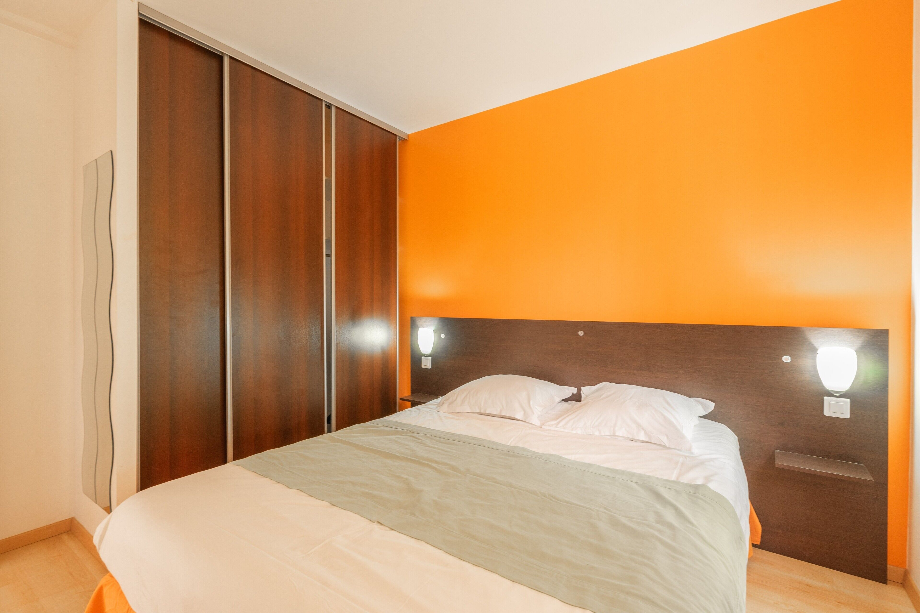 A bedroom with a bed, a wooden wardrobe, and a wall-mounted light fixture.