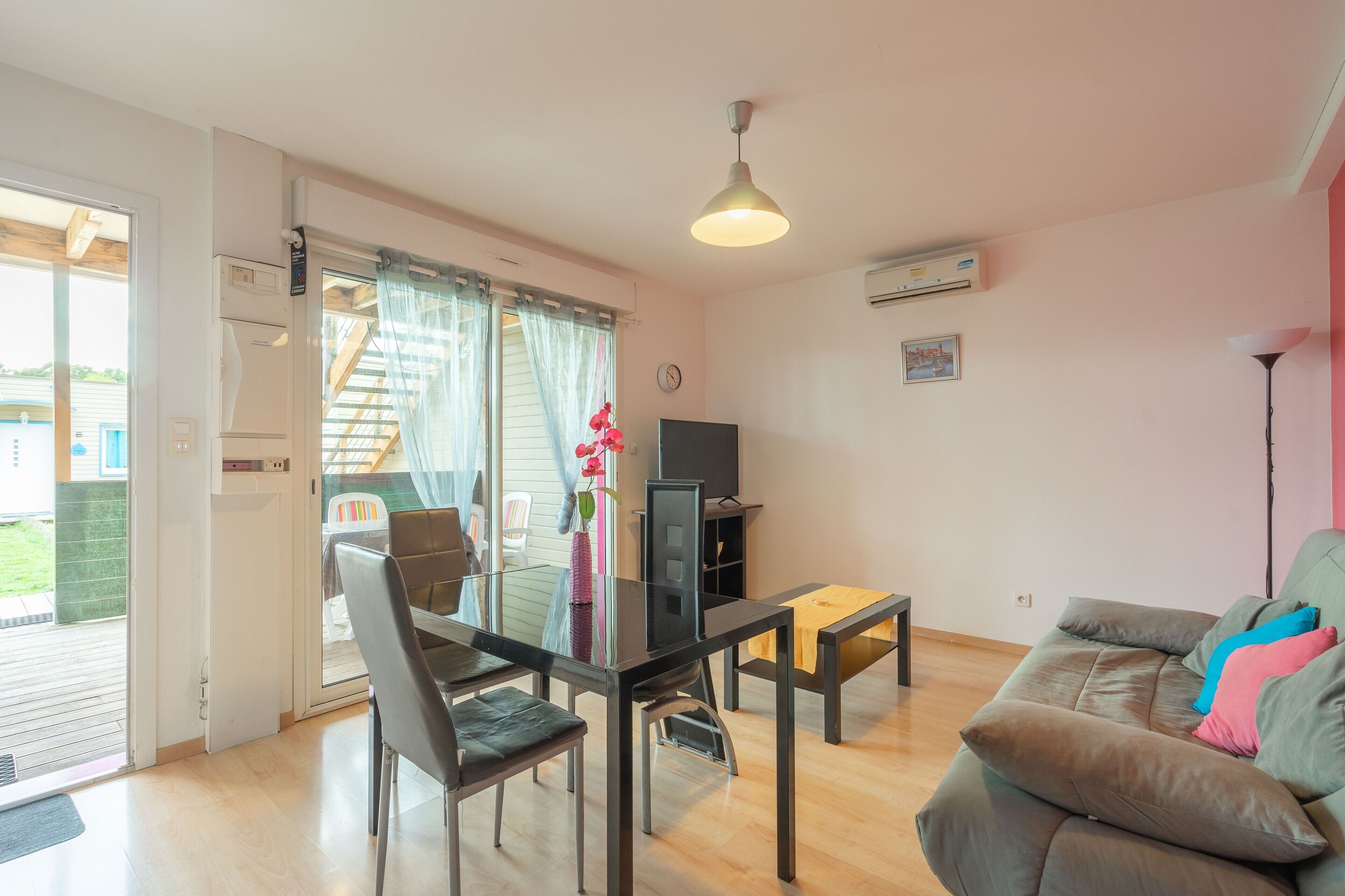 A living room with a sofa, a TV, a table, and a chair. There is a door leading to an outdoor area.
