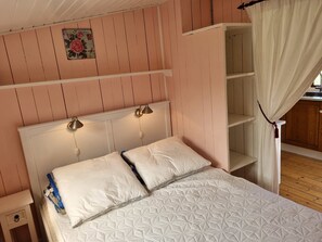 A bedroom with a bed, bedside lamps, a nightstand, a shelf, and a curtain. - Charming and cozy house near Høve beach (Asnæs)