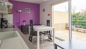 Eine moderne Küche mit weißer Arbeitsplatte, einer Kaffeemaschine und einer Vase mit Blumen. Ein Esstisch mit Stühlen und ein großes Fenster mit Blick auf eine Holzterrasse und Grünflächen.
