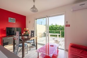 Un salon moderne avec un mur rouge, un téléviseur à écran plat, une table à manger avec des chaises, un canapé rouge et une porte-fenêtre coulissante donnant sur une terrasse extérieure.