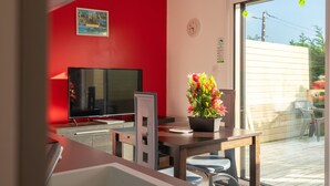 Ein Zimmer mit roter Wand, einem Fernseher, einem Esstisch mit Stühlen und einem Fenster mit Blick ins Freie.
