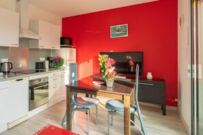 Une cuisine moderne avec des meubles blancs, un mur d’accent rouge, un coin repas avec une table et des chaises, et un téléviseur.